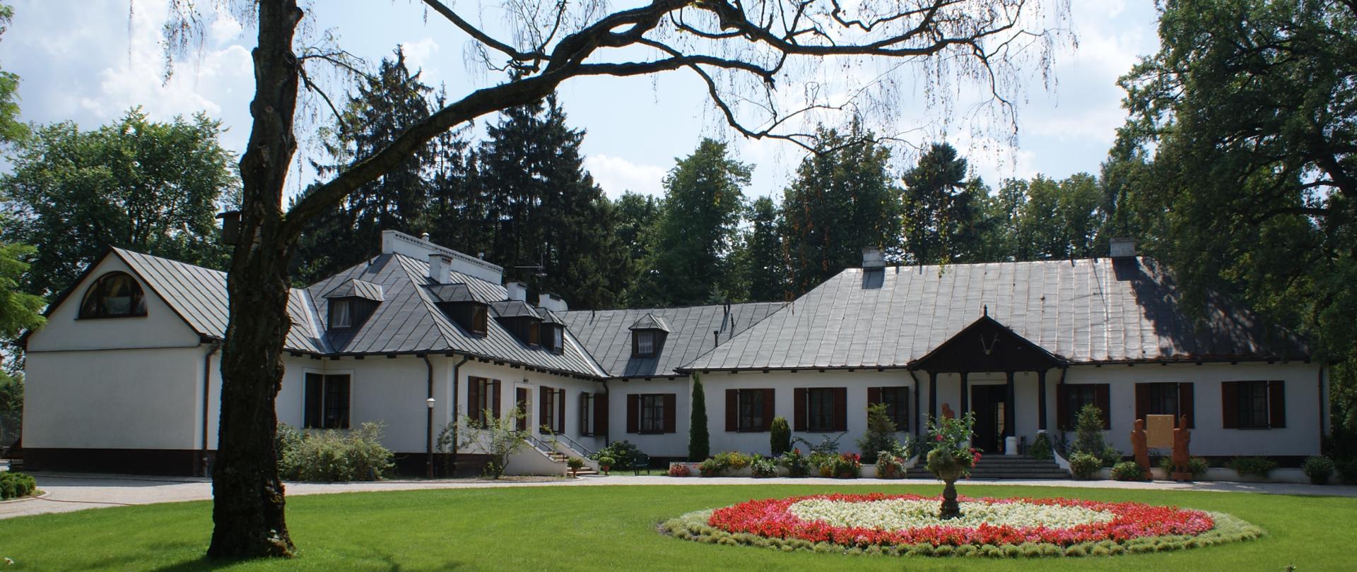 Grafika przedstawia zdjęcie Domu Pracy Twórczej „Reymontówka” w Chlewiskach. Biały dworek z czarnym dachem, nad wejściem ganek z czterema kolumienkami i dwuspadowym czarnym dachem.