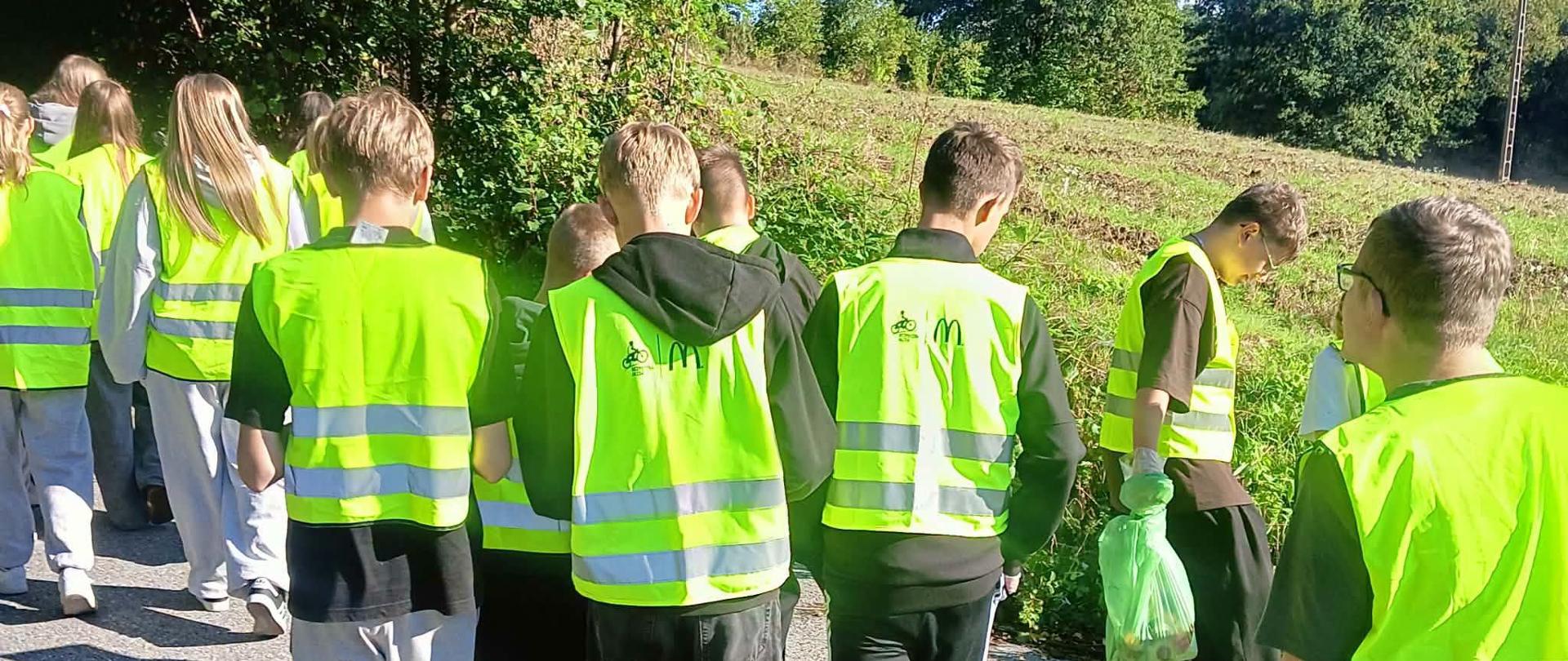 Na zdjęciu widać grupę nastolatków ubranych w jaskrawożółte kamizelki odblaskowe. Idą poboczem drogi wzdłuż pól i łąk. Kilka osób trzyma duży czarny worek, prawdopodobnie podczas zbierania odpadów. W tle i na krzewach widać liście z oznakami jesieni.