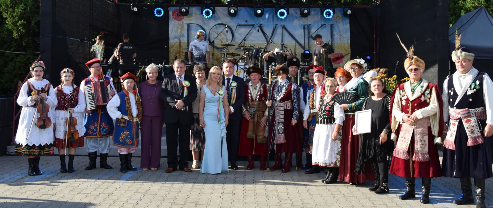 Duża grupa ludzi pozuje do zdjęcia na brukowanym placu przed sceną. Niektórzy są ubrani w tradycyjne polskie stroje ludowe, inni w stroje szlacheckie (kontusze), a jeszcze inni w garnitury i sukienki bankietowe. W tle widać scenę z instrumentami muzycznymi i napisem „DOŻYNKI”.