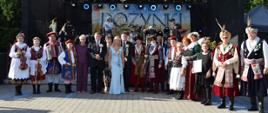 Duża grupa ludzi pozuje do zdjęcia na brukowanym placu przed sceną. Niektórzy są ubrani w tradycyjne polskie stroje ludowe, inni w stroje szlacheckie (kontusze), a jeszcze inni w garnitury i sukienki bankietowe. W tle widać scenę z instrumentami muzycznymi i napisem „DOŻYNKI”.