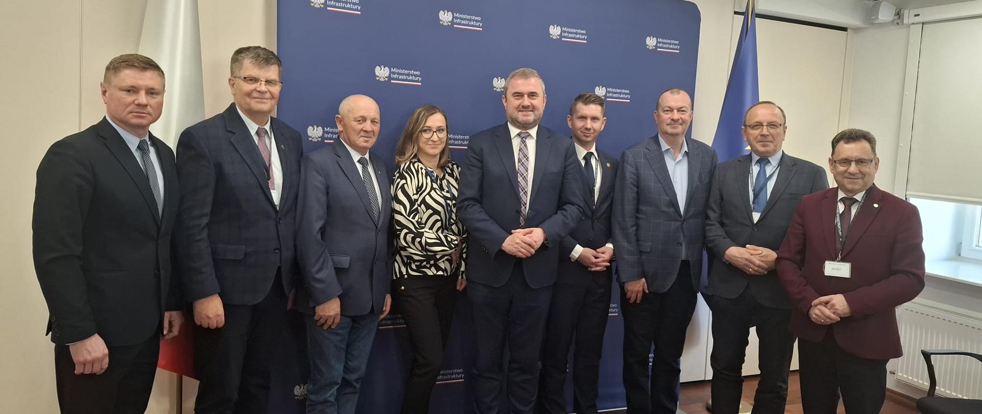 Zdjęcie grupowe na tle granatowej ścianki z Ministerstwo Infrastruktury. Na zdjęciu uwieczniono osoby wraz wiceministrem. W tle flaga Polski ora Unii Europejskiej.