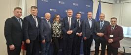 Zdjęcie grupowe na tle granatowej ścianki z Ministerstwo Infrastruktury. Na zdjęciu uwieczniono osoby wraz wiceministrem. W tle flaga Polski ora Unii Europejskiej.