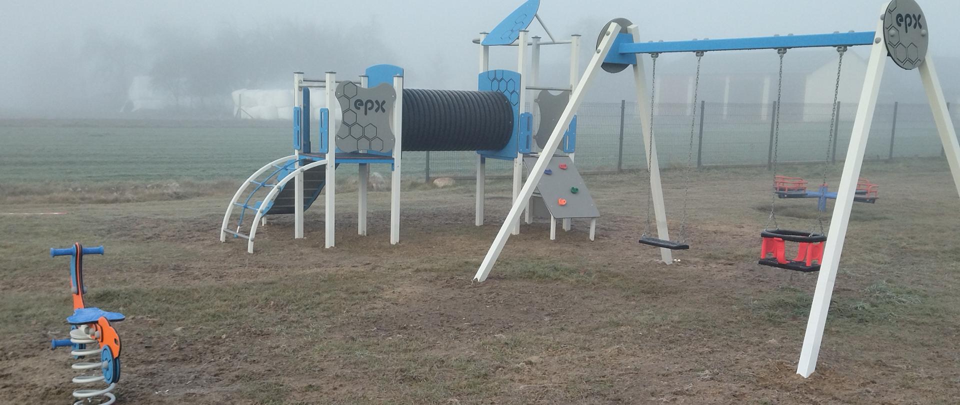 Obraz przedstawia plac zabaw na otwartym terenie, częściowo otoczony ogrodzeniem. W centralnej części znajduje się huśtawka z dwoma siedziskami, wsparta na metalowej konstrukcji w kolorze niebieskim. Po lewej stronie widoczny jest bujak sprężynowy w kształcie motocykla, pomalowany na czerwono i żółto. Na placu zabaw znajduje się także zestaw do wspinaczki, zawierający tunel oraz różne elementy do zabawy.