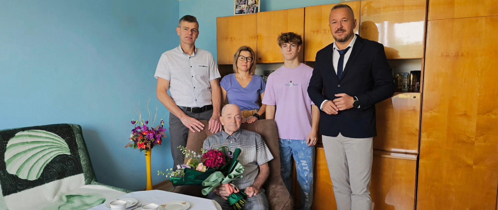 Grupa osób pozuje do zdjęcia ze starszym mężczyzną siedzącym na fotelu z bukietem kwiatów