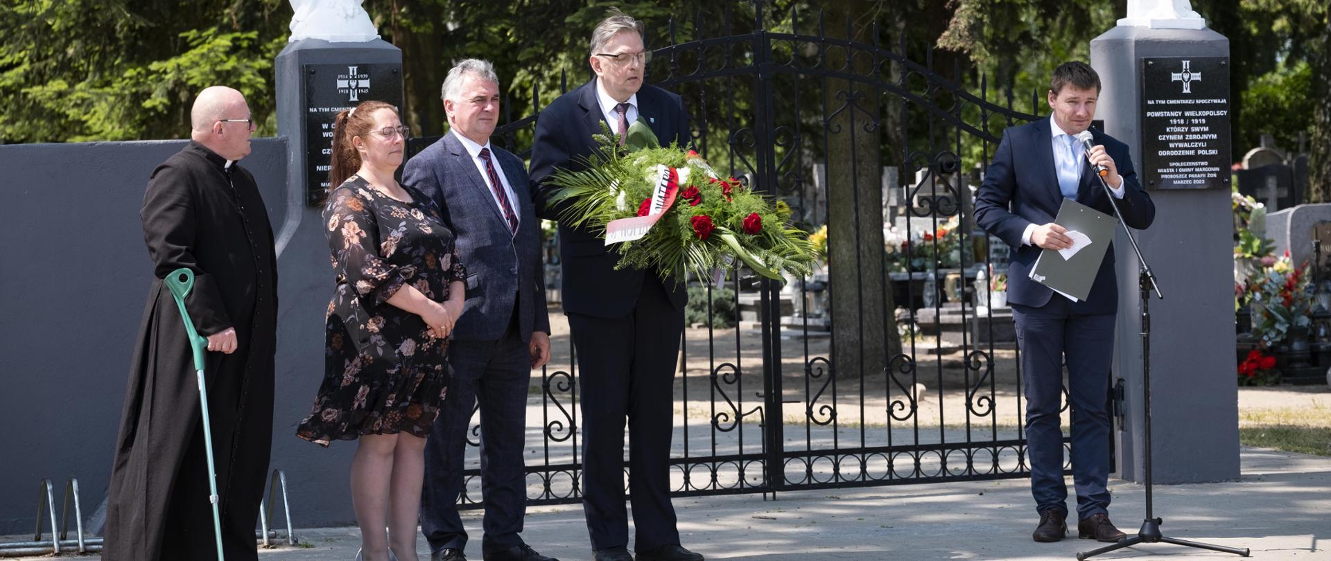 Ksiądz, kobieta, burmistrz Margonina, przewodniczący Rady Miasta i Gminy Margonin oraz zastępca burmistrza Margonina stojący na tle bramy wjazdowej cmentarza w Żoniu. Zastępca Burmistrza przemawia do mikrofonu na statywie.