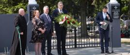Ksiądz, kobieta, burmistrz Margonina, przewodniczący Rady Miasta i Gminy Margonin oraz zastępca burmistrza Margonina stojący na tle bramy wjazdowej cmentarza w Żoniu. Zastępca Burmistrza przemawia do mikrofonu na statywie.