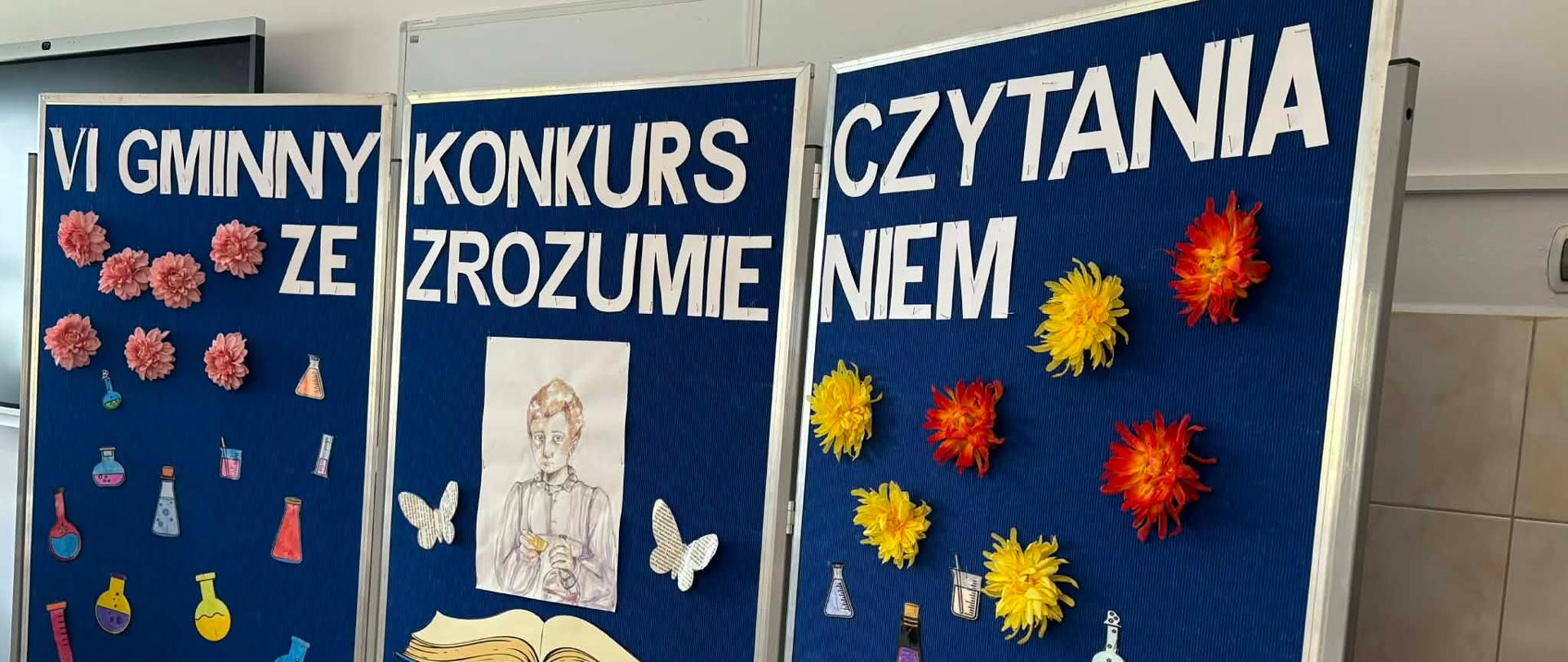 Gablota o niebieskim kolorze na którym widnieje biały napis. Poniżej napisu przywieszona jest dekoracja- książka, akcesoria chemiczne oraz porter - Marii Curie - Skłodowskiej