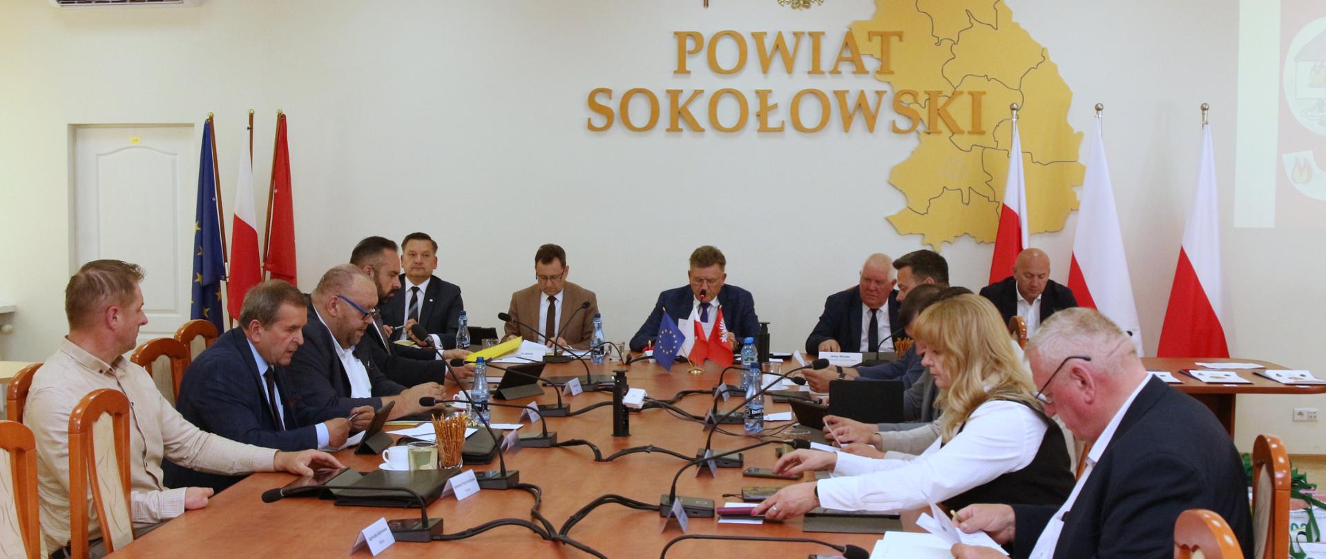 Fotografia wykonana podczas XIII Sesji Rady Powiatu Sokołowskiego. Trwają obrady, przy stole na sali konferencyjnej siedzą radni, natomiast na wprost nich siedzi wicestarosta, starosta, przewodniczący rady powiatu oraz dwóch wiceprzewodniczących. Na ścianie napis powiat sokołowski, krzyż, godło Polski oraz wycina obszaru powiatu sokołowskiego.
