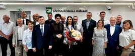Zdjęcie grupowe po obradach sesji na tle ściany z logiem i napisem "GMINA KOBIELE WIELKIE", w centralnym miejscu Wójt Gminy Kobiele Wielkie Beata Pokora z kwiatami, po lewej stronie Skarbnik Anna Czerkiewska oraz Zastępca Wójta Andrzej Kuna; po lewej i prawej stronie wszyscy 15 Radnych Rady Gminy Kobiele Wielkie. 