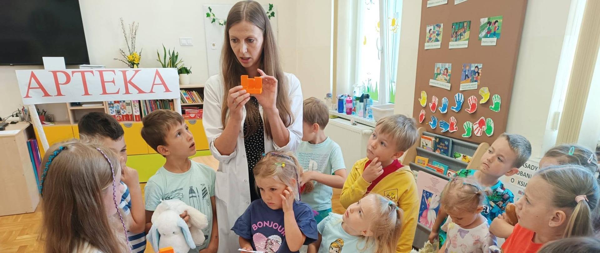 Na zdjęciu widać grupę dzieci zgromadzonych wokół stołu podczas zajęć edukacyjnych w sali przedszkolnej lub szkolnej. W centrum stoi kobieta ubrana w biały fartuch, która prezentuje dzieciom pomarańczowy pojemnik przypominający opakowanie na leki. Obok niej znajduje się druga osoba również w białym fartuchu, co sugeruje, że zajęcia dotyczą tematyki zdrowia, farmacji lub pierwszej pomocy.
Na stole rozłożone są różne przybory i materiały edukacyjne, m.in. pojemniki, buteleczki, moździerz, dzbanek oraz karty pracy. Nad stanowiskiem widnieje duży napis „APTEKA”, wskazujący na temat zajęć. Dzieci z dużym zainteresowaniem słuchają prowadzącej, obserwując prezentowane przedmioty i zadając pytania.
Sala jest jasna i kolorowa. W tle znajdują się książki, dekoracje oraz tablica z pracami plastycznymi dzieci. Atmosfera na zdjęciu jest pełna ciekawości i zaangażowania, a zajęcia mają charakter praktyczny i edukacyjny, pozwalając dzieciom poznawać pracę farmaceuty oraz zasady dbania o zdrowie.