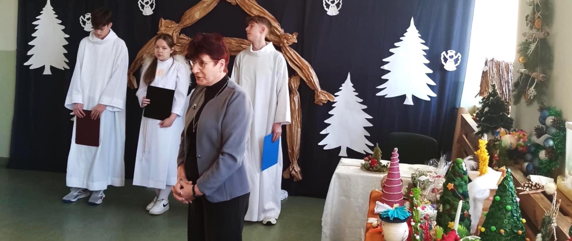 Na fotografii na pierwszym planie dyrektor Niepublicznej Szkoły Podstawowej im. Armii Krajowej w Niecieczy przemawia podczas uroczystego rozstrzygnięcia powiatowego konkursu na ozdobę bożonarodzeniową/ W tle trzech uczniów przebranych na biało.