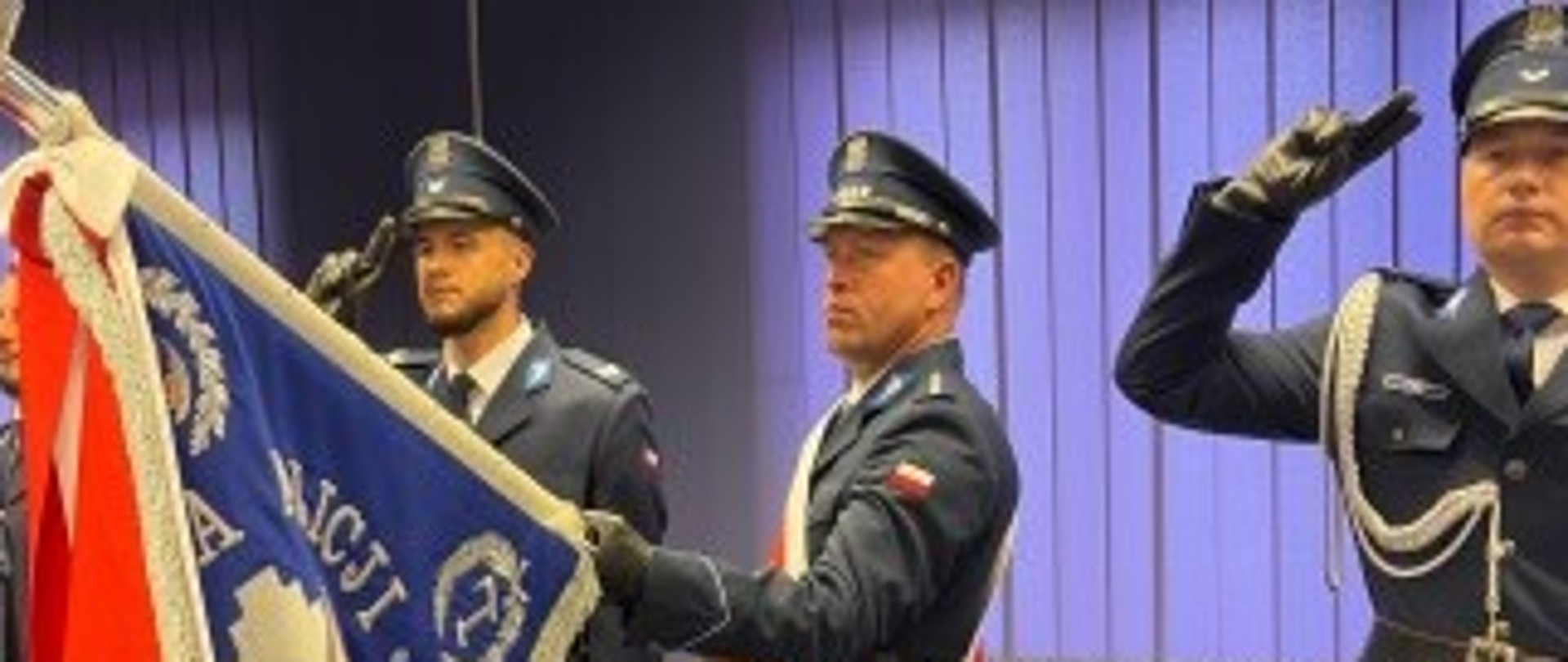 grupa osób w mundurach galowych policji. W tym poczet sztandarowy. Jeden mężczyzna klęczy, jeden salutuje.