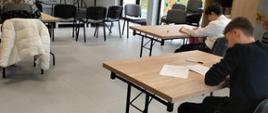 Jedna uczennica i dwóch uczniów siedzą pojedynczo przy stolikach i piszą arkusz konkursowy, przestronna jasna sala z dużymi oknami, czarne krzesła, drewniane szafki