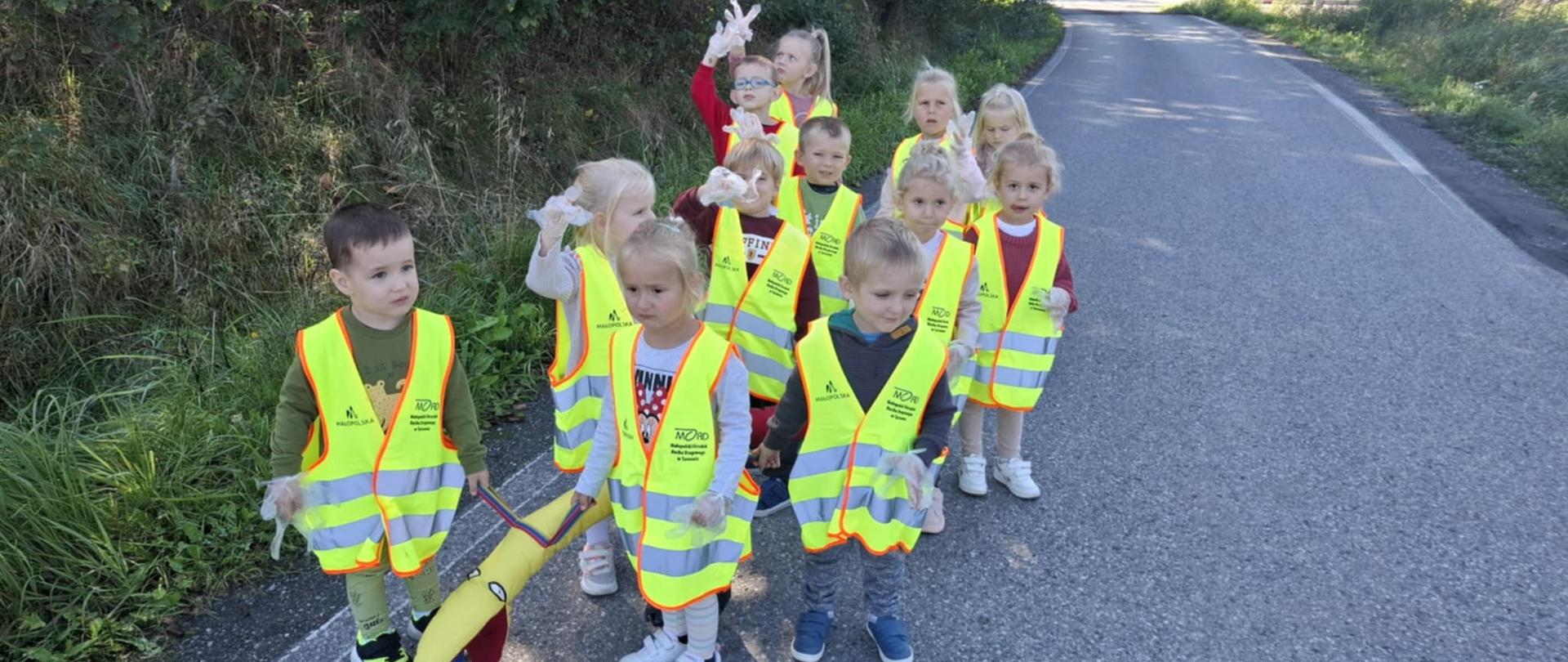 Grupa dzieci w parach idzie drogą w kamizelkach odblaskowych i rękawiczkach jednorazowych.