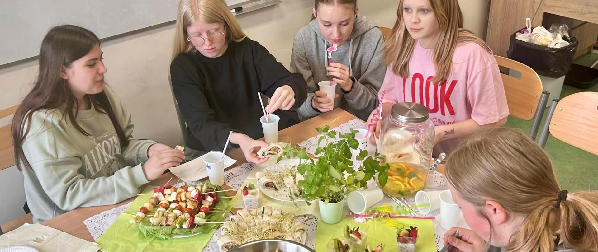 Pięć dziewczynek siedzi przy stole na którym są serwetki białe i limonowe, talerze, sztuce, kolorowe szaszłyki owocowe, sałatka warzywna, zdrowe przekąski warzywno-owocowe.