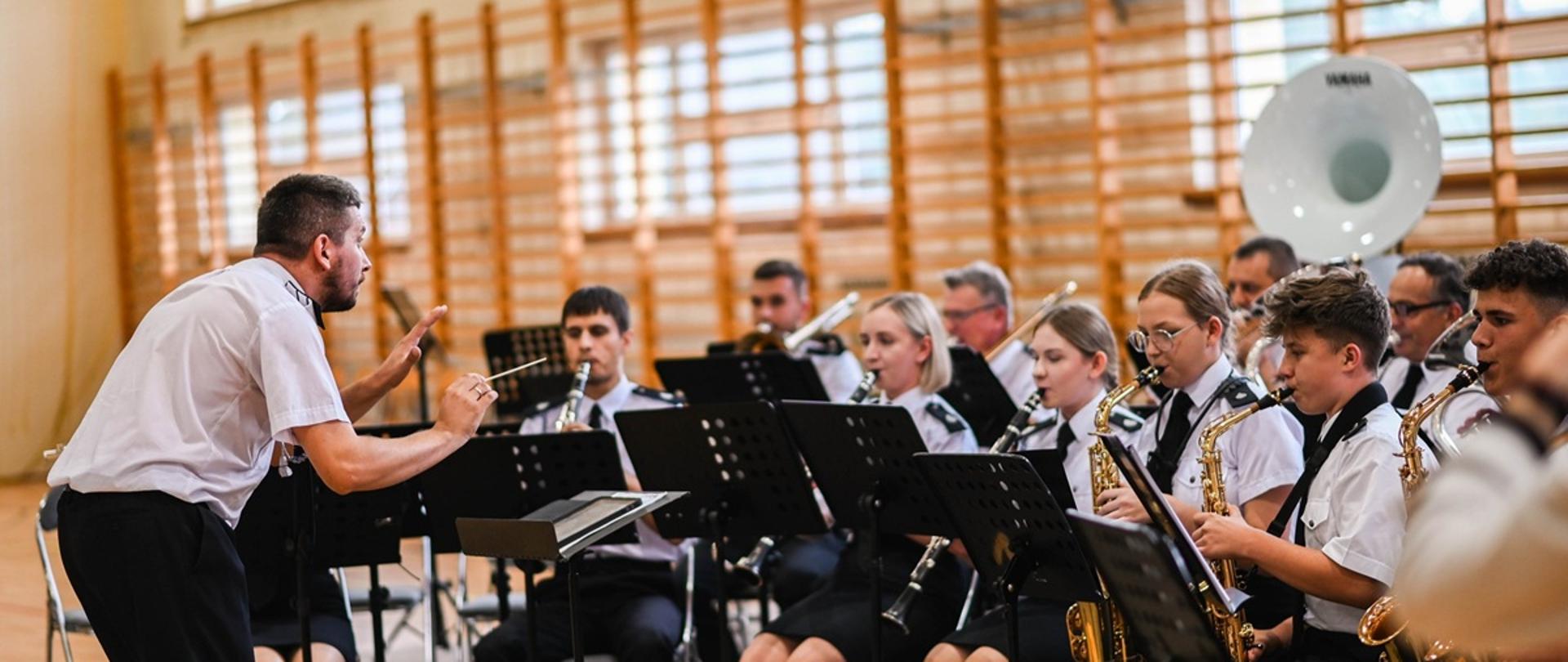 członkowie orkiestry dętej siedzą na krzesłach i grają na instrumentach, dyrygent nachylony do nich dyryguje 