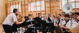 członkowie orkiestry dętej siedzą na krzesłach i grają na instrumentach, dyrygent nachylony do nich dyryguje 