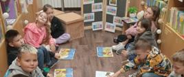Dzieci z klasy 3 siedzą na piankowych matach w bibliotece w kręgu naprzeciw siebie, każdy z nich czyta głośno po fragmencie książki, która leży przed nimi na podłodze, po lewej i prawej stronie za dziećmi półki z książkami