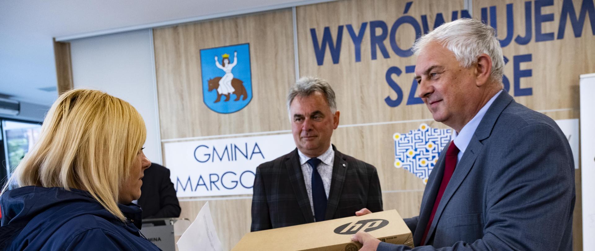 Minister Piechowiak wręczający 2 komputery przenośne kobiecie, w tle burmistrz Janusz Piechocki, za nim ściana z herbem Gminy Margonin i napisem Wyrównujemy szanse