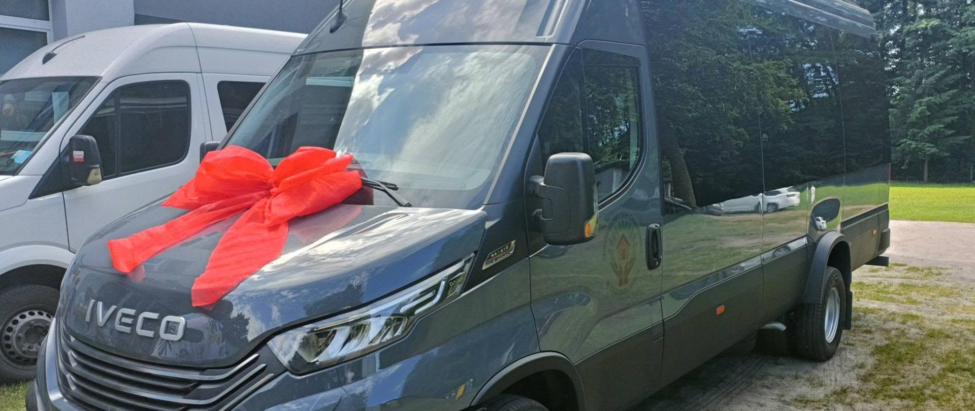 Zdjęcie prezentuje profil nowego, ciemnoszarego busa marki Iveco. Pojazd posiada przyciemniane szyby oraz duże ozdobne czerwone kokardy umieszczone na przedniej masce. Auto stoi na piaszczystym terenie, w tle widoczny jest fragment budynku oraz zieleń. Na przednim zderzaku widoczna jest tablica rejestracyjna o numerze EPA 60999. Na boku pojazdu znajduje się słabo widoczne logo instytucji.