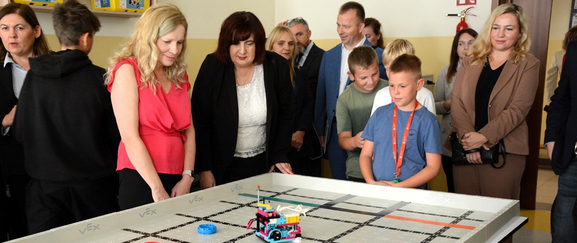 mężczyźni i kobiety stoją wokół stołu na którym jeździ robot zbudowany z klocków lego 