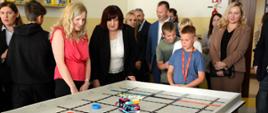 mężczyźni i kobiety stoją wokół stołu na którym jeździ robot zbudowany z klocków lego 