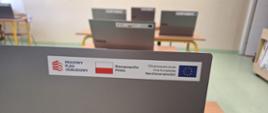 Zbliżenie na srebrną obudowę laptopa na naklejką, na której widnieje od lewej: logo Krajowego Planu Odbudowy, flaga Polski biało-czerwona oraz flaga Unii Europejskiej