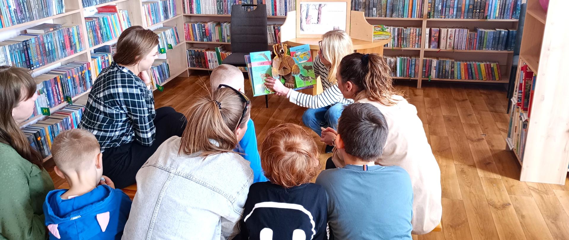 Zdjęcie przedstawia dzieci przedszkolne wraz z paniami oglądające prezentowaną przez bibliotekarkę książkę z wychodzącym z niej papierowym niedźwiedziem