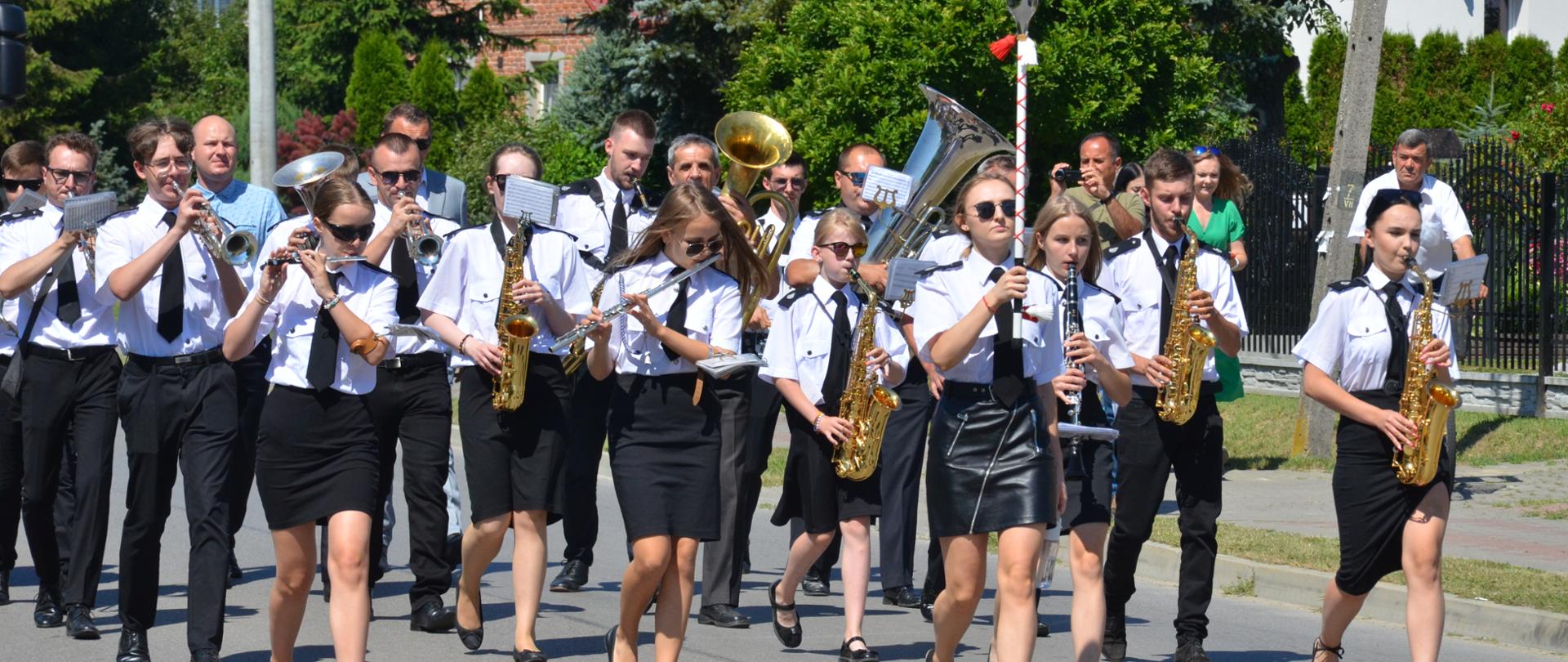 Grupa muzykantów grająca na różnych instrumentach muzycznych maszerując po środku drogi