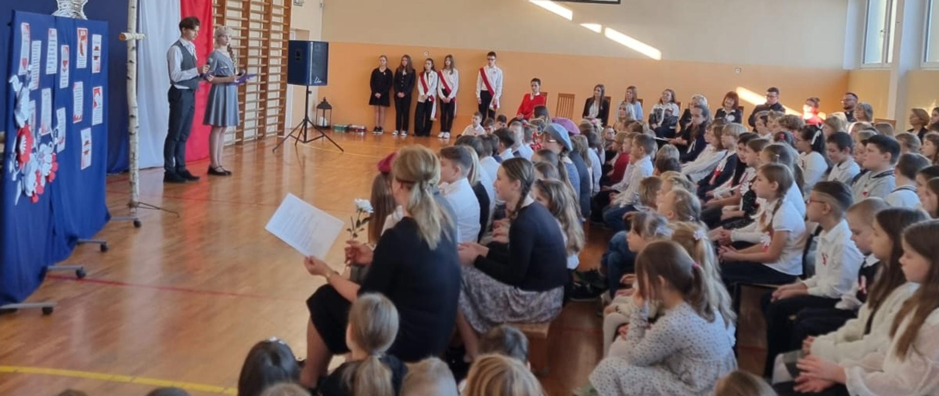 Społeczność szkolna ogląda występ uczniów z okazji Narodowego Święta Niepodległości w sali gimnastycznej