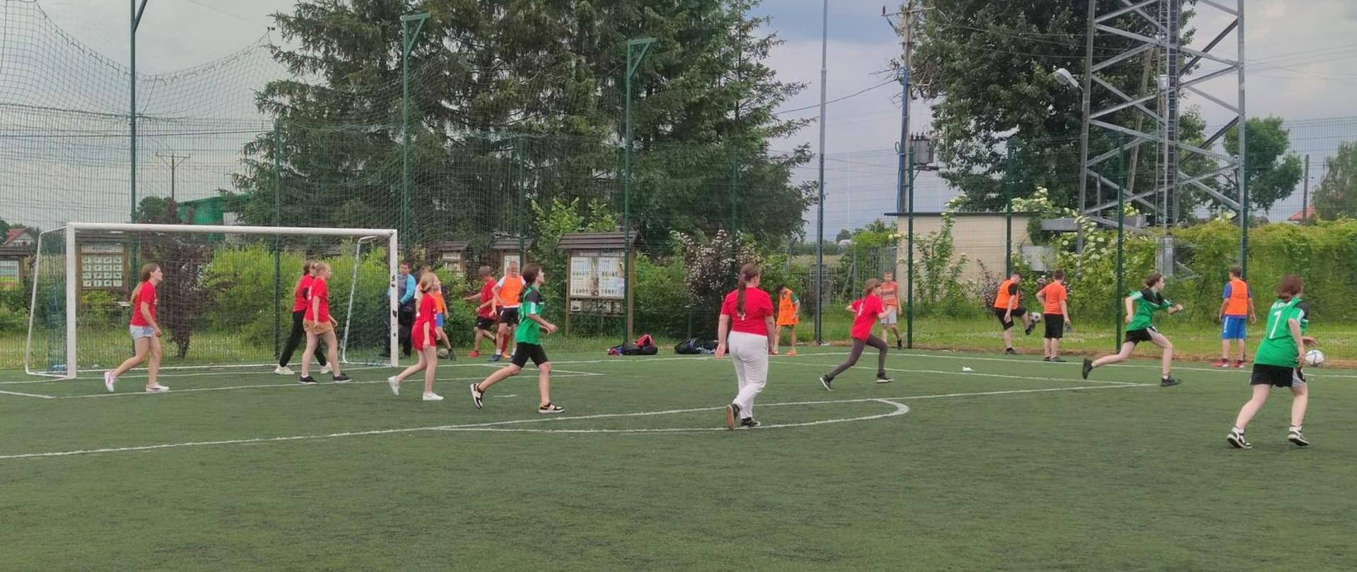 Dwie drużyny dziewcząt grające w piłkę nożną, jedna drużyna w czerwonych koszulkach, druga w zielonych koszulkach i czarnych spodenkach.