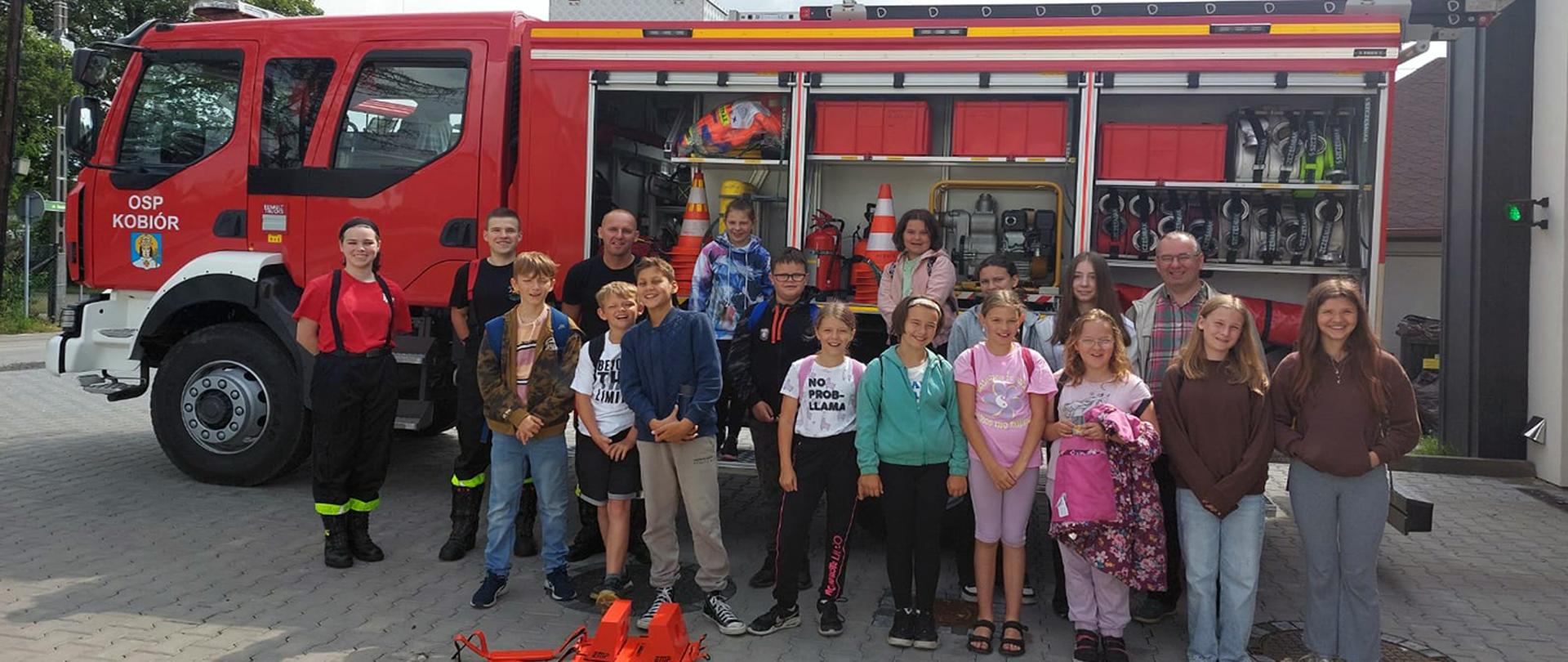 Grupa dzieci wraz z opiekunem oraz strażakami OSP na tle wozu strażackiego