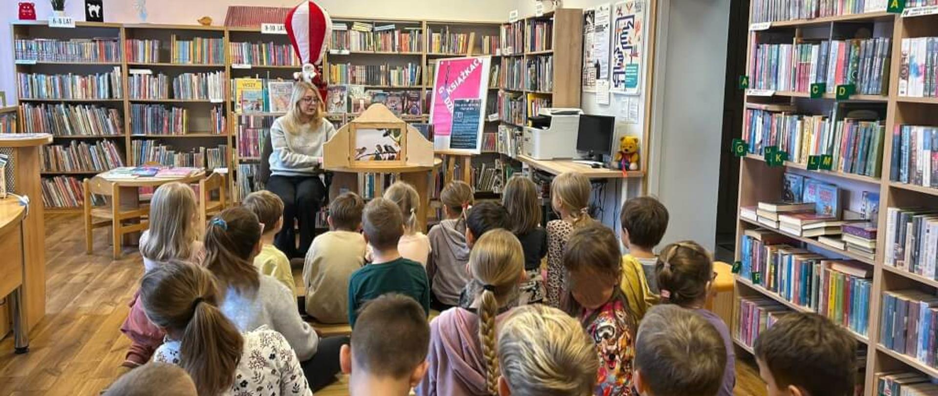 Na zdjęciu widzimy grupę dzieci, którzy słuchają opowieści Pani prowadzącej zajęcia biblioteczne dla dzieci. Pani Bibliotekarka opowiadania bajkę „Jak przetrwasz mroźną zimę kopciuszku”, które porusza temat migracji ptaków.