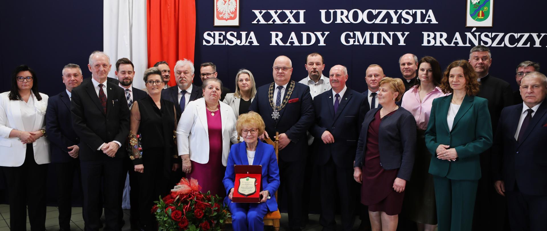 Wnętrze sali osp w Brańszczyku, na wspólnym zdjęciu uczestnicy wydarzenia, starsza kobieta siedzi na krześle z grawertonem, obok kosz z różami.