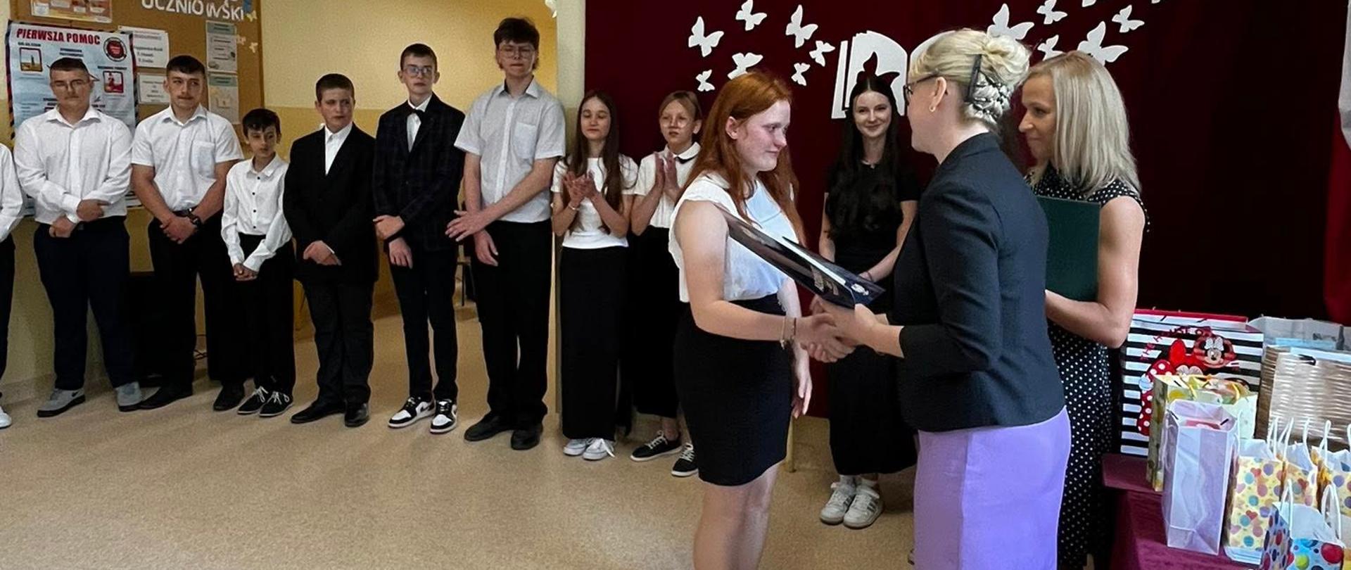 Pani dyrektor wręcza świadectwo i nagrodę uczennicy klasy 8, obok stoi wychowawczyni i pozostali uczniowie klasy 8, na ścianie na bordowej kotarze umieszczona dekoracja w postaci motylków unoszących się znad otwartej książki i napis "Pożegnania nadszedł czas..."