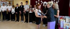 Pani dyrektor wręcza świadectwo i nagrodę uczennicy klasy 8, obok stoi wychowawczyni i pozostali uczniowie klasy 8, na ścianie na bordowej kotarze umieszczona dekoracja w postaci motylków unoszących się znad otwartej książki i napis "Pożegnania nadszedł czas..."