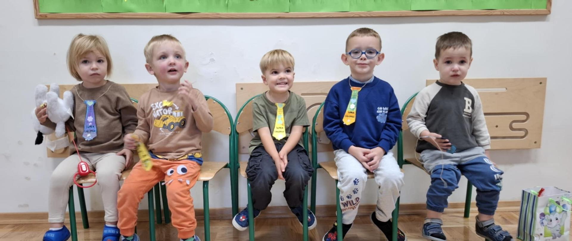 Pięciu małych chłopców siedzi na krzesełkach w sali pod ścianą. Wszyscy uśmiechnięci i z kolorowymi papierowymi krawatami na szyi.