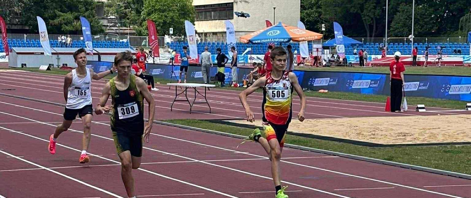 Troje chłopców ubranych w stroje sportowe z numerami na koszulkach biegnie po tartanowej bieżni.