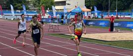 Troje chłopców ubranych w stroje sportowe z numerami na koszulkach biegnie po tartanowej bieżni.