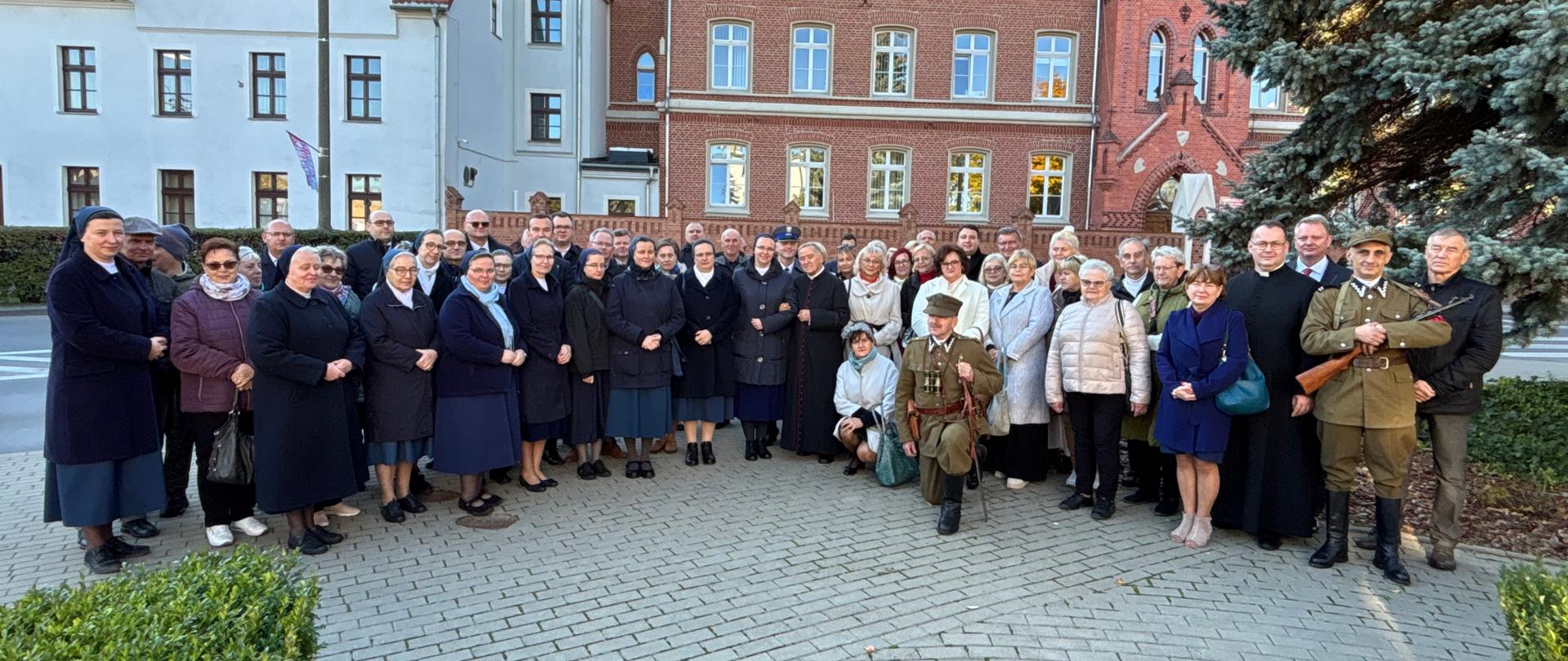 Grupa osób uczestników uroczystości (ponad 50 osób) stoi na chodniku z jasnej, szarej kostki brukowej. W tle dwa budynki, jeden o białej elewacji z dachem pokrytym czerwoną dachówką, drugi cały z czerwonej cegły. 