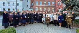 Grupa osób uczestników uroczystości (ponad 50 osób) stoi na chodniku z jasnej, szarej kostki brukowej. W tle dwa budynki, jeden o białej elewacji z dachem pokrytym czerwoną dachówką, drugi cały z czerwonej cegły. 