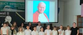 Na scenie znajduje się kilkunastu uczniów biorących udział w apelu. Jedna z dziewczynek przemawia przed mikrofon. Wszyscy ubrani są na galowo. Z tyłu wyświetlona jest fotografia Jana Pawła II. 