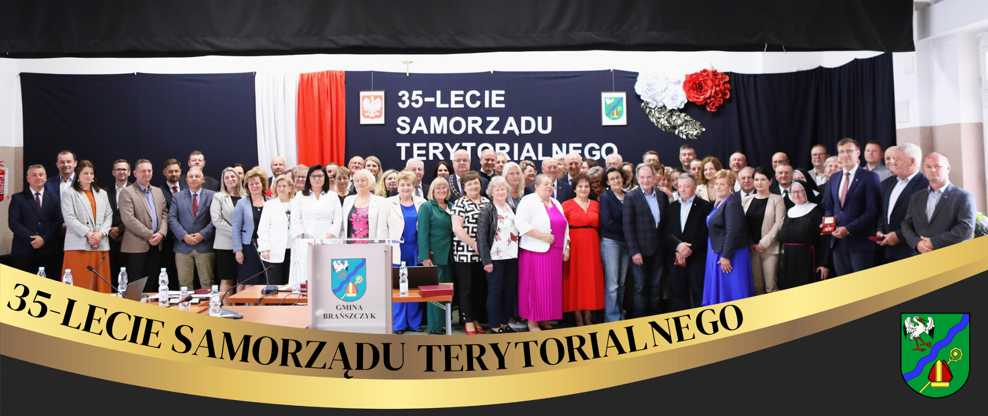 Wnętrze sali osp, na tle dekoracji grupa kilkudziesięciu osób pozuje do zdjęcia, poniżej ozdobna fala z napisem 35-lecie samorządu terytorialnego i herb gminy