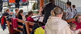 Zasłużony odpoczynek po akcji sprzątania ziemi. Dzieci oraz opiekunowie siedzą pod drewnianą wiatą przy drewnianych stolikach i jedzą pizzę. 