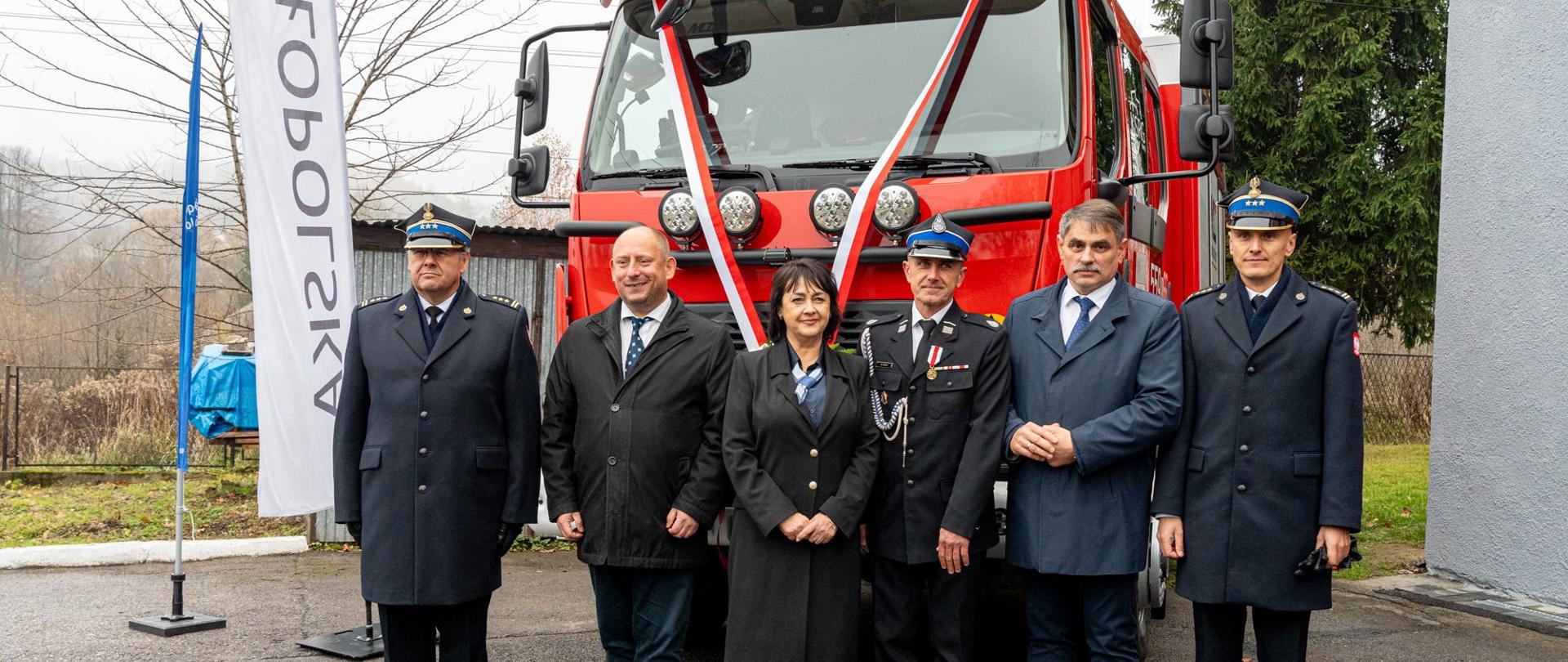 Na zdjęciu stoi sześć osób, ustawionych przed nowym czerwonym samochodem ratowniczo-gaśniczym udekorowanym biało-czerwoną wstęgą. Wśród obecnych są funkcjonariusze straży pożarnej w galowych mundurach oraz przedstawiciele władz samorządowych, w tym kobieta i kilku mężczyzn w eleganckich płaszczach. Po lewej stronie widoczne są flagi z napisem „Małopolska”. Tło stanowi teren przy remizie OSP – drzewo bez liści, fragment ogrodzenia oraz zabudowania gospodarcze.