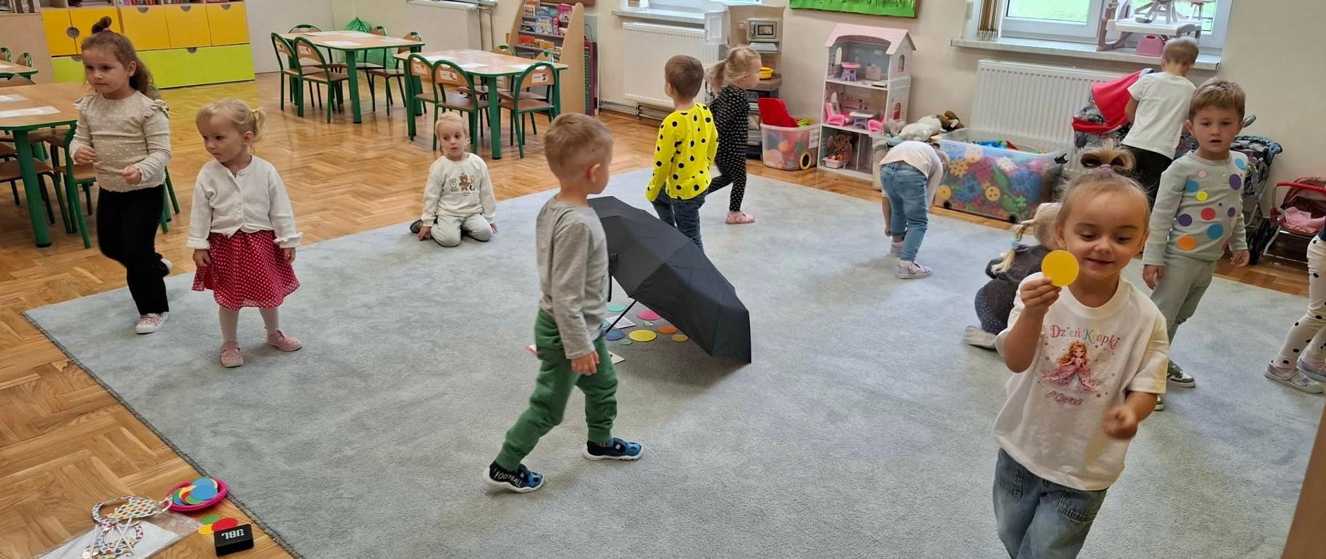 Na zdjęciu widać salę przedszkolną, w której bawi się grupa małych dzieci. Pomieszczenie jest jasne i przestronne – znajdują się w nim stoliki z krzesełkami, półki z książkami i zabawkami oraz duży szary dywan pośrodku sali. Dzieci wykonują różne czynności: chodzą, schylają się po przedmioty, bawią się zabawkami i kolorowymi krążkami. Jedna dziewczynka na pierwszym planie trzyma w ręce żółte kółko i uśmiecha się do aparatu. Na dywanie leży również otwarty parasol, który prawdopodobnie jest elementem zabawy lub zajęć. W tle można zauważyć domek dla lalek, pojemniki z zabawkami oraz dekoracje na ścianach przedstawiające jesienne motywy, takie jak drzewo i grzyby. Za oknami widoczny jest plac zabaw. Atmosfera na zdjęciu jest radosna i pełna dziecięcej aktywności.