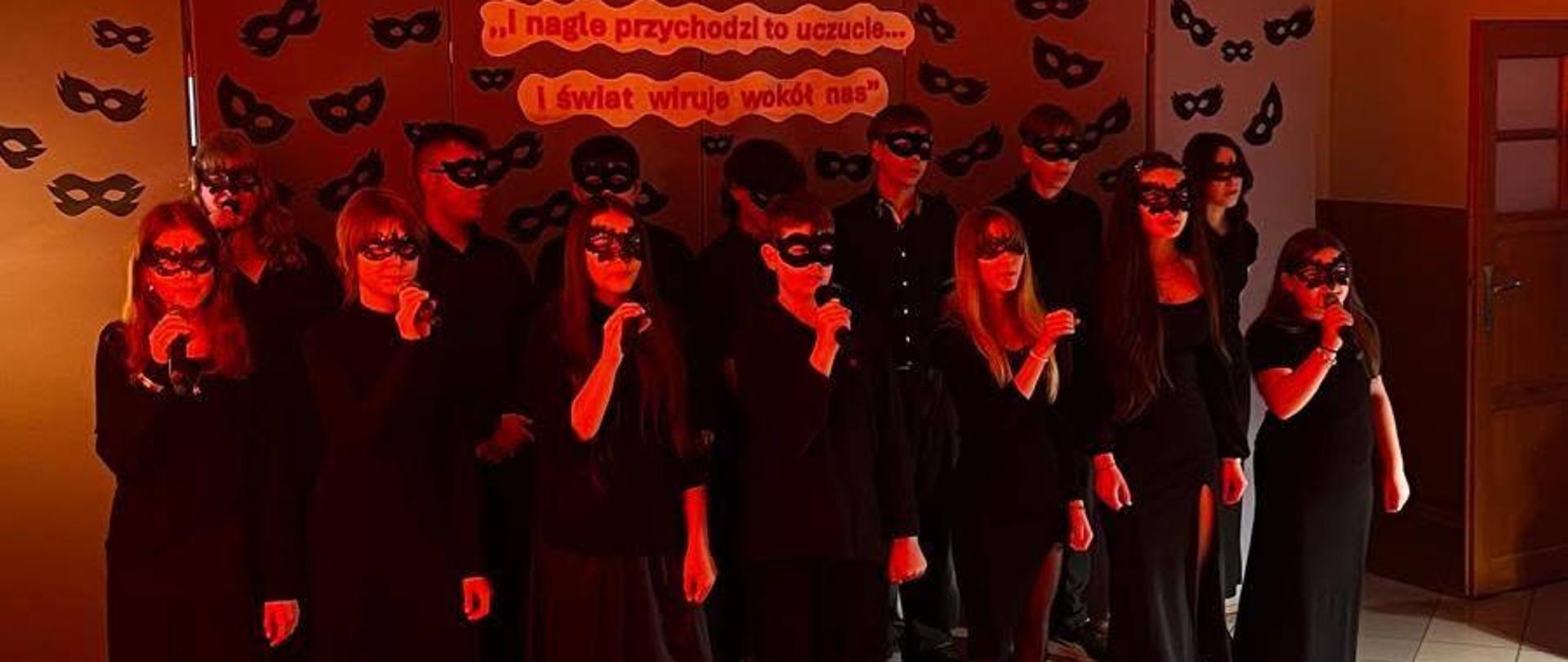 Zdjęcie przedstawia grupę młodzieży ubraną na czarno i w maskach karnawałowych, stojącą w rzędach na korytarzu szkolnym lub sali, oświetloną intensywnym, czerwonym światłem.