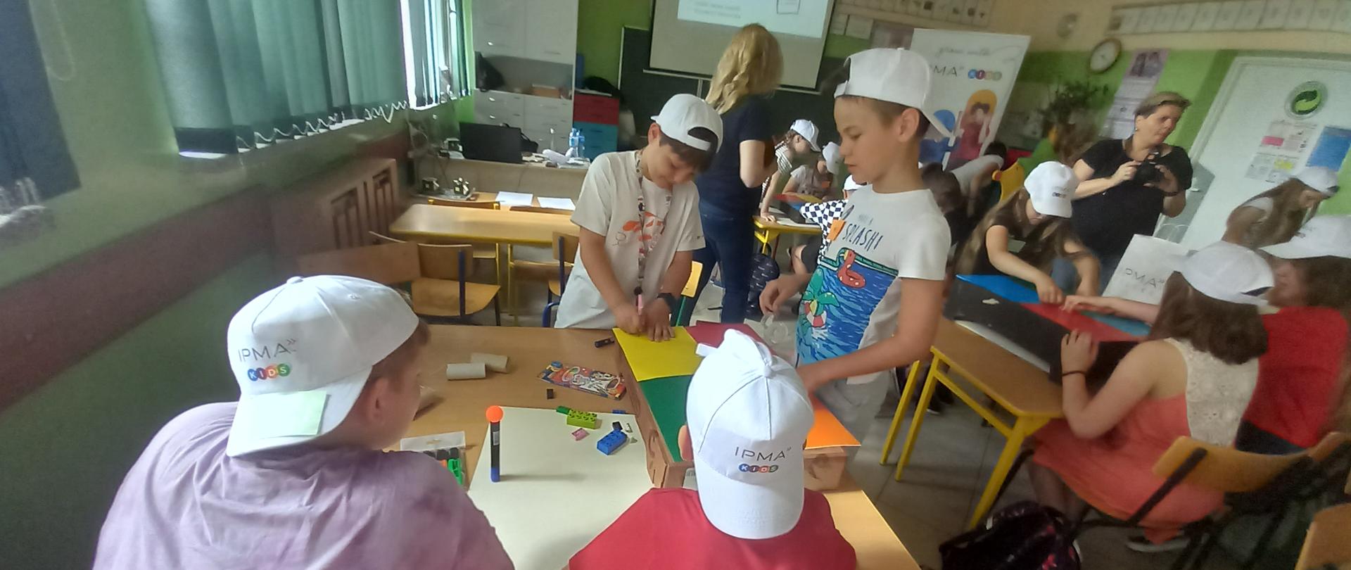 Dzieci siedzą przy ławkach i zapisują coś na kolorowych kartkach