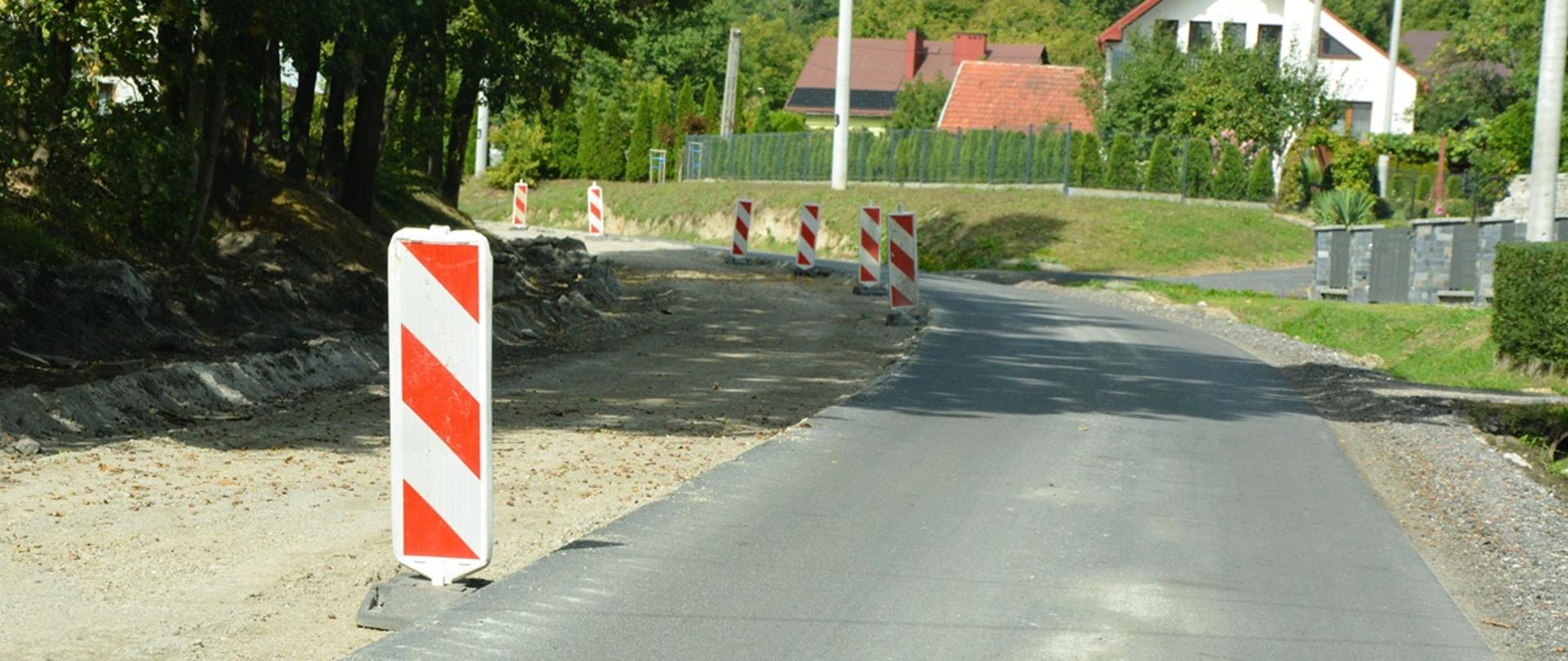 zdjęcie przedstawia jezdnie w remoncie, po lewej stronie brak jezdni, widoczny jest piasek i kamienie po prawej normalny pas jezdni, w środku słupki biało czerwone w tle zielone drzewa i domy 