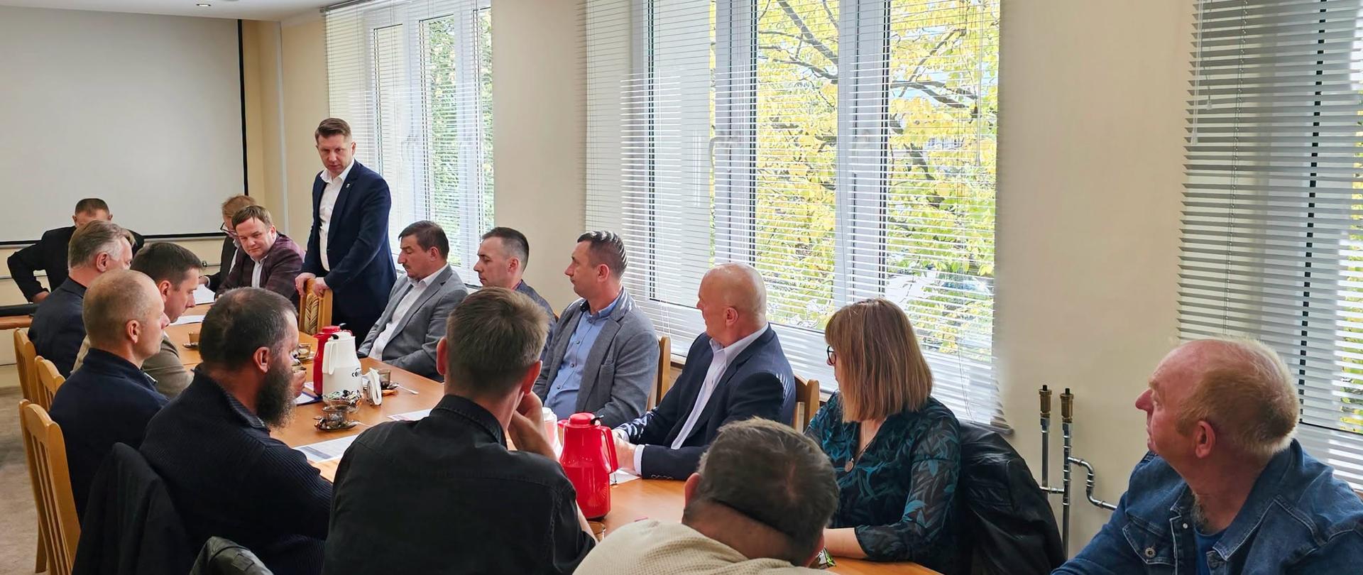 Grupa osób siedzących przy stole konferencyjnym, prowadząca dyskusję z kilkoma osobami stojącymi i przedstawiającymi prezentację. Duże okna w tle wpuszczają naturalne światło.